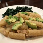 pasta with white bean sauce topped with sliced avocado with a bed of kale in the background