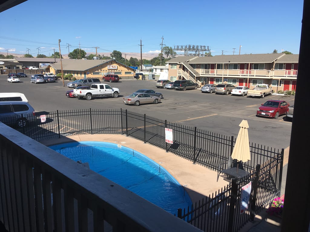 View outside the motel room I stayed in. A small pool in the foreground surrounded by a black metal gate. A large parking lot with another motel building and a Mexican restaurant in the background.