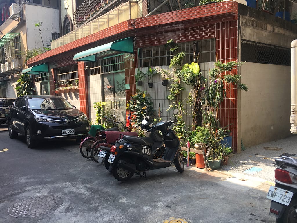 Alley in Taipei. Four parked scooters in the foreground, one very sleek car, and older buildings covered in red tile with plants growing up the walls.