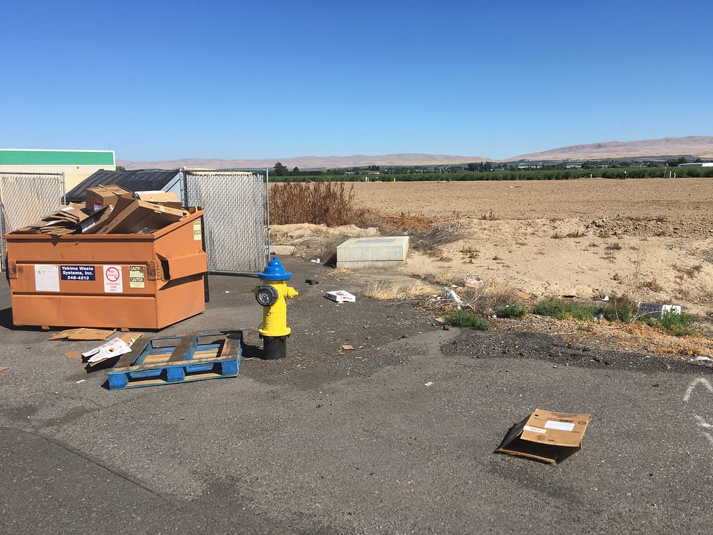 Orange Dumpster overflowing with cardboard boxes next to a discarded wooden pallet and a bright yellow fire hydrant, with brown flat land behind them, and a stripe of green farmland and dusty brown hills in the distance.