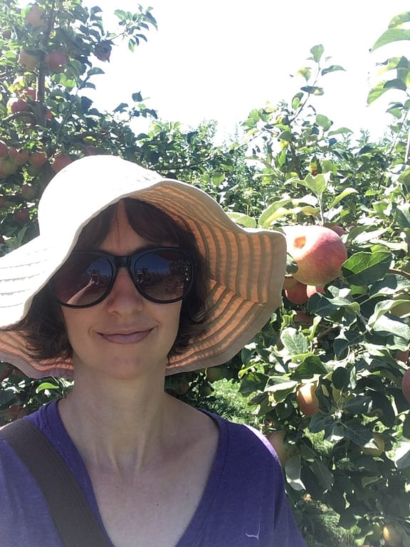 The author in a big floppy sun hat in sunglasses, surrounded by apple trees