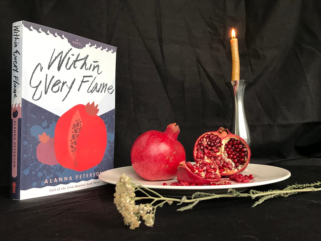 The paperback edition of Within Every Flame stands in front of a black backdrop, surrounded by pomegranates, a sprig of dried yarrow, and a burning beeswax candle inside a tall silver candleholder.