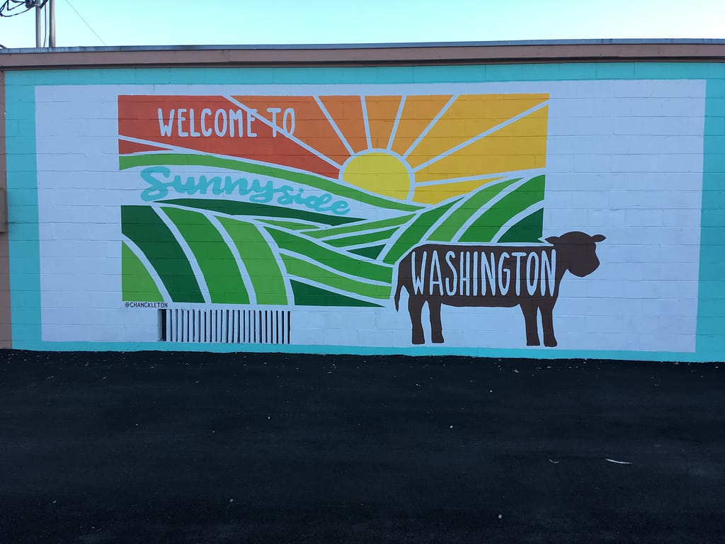 Mural painted on the side of a building, showing green rolling pastures, a sunny sky, a silhouette of a cow, and the words, "Welcome to Sunnyside Washington."