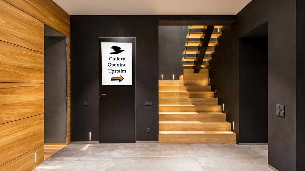 the lobby of a fancy looking gallery with black walls and light wood staircase. a sign on the wall reads "gallery opening upstairs" with an arrow pointing towards the staircase