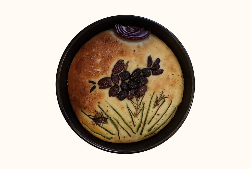 A circular loaf of focaccia inside a pan sitting on a shelf. A crow made of olives flies across the center. A red-onion sun and chive grass round out the composition.