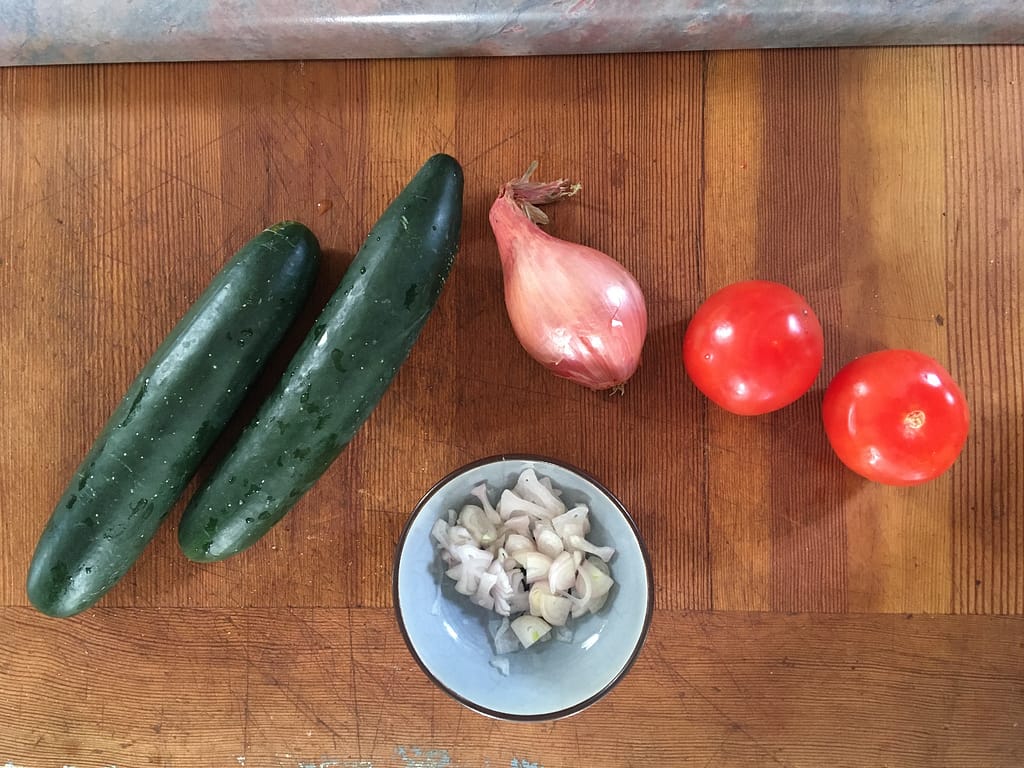 whole cucumbers, shallots, and tomatoes on a wooden cutting board, plus a small bowl of chopped shallots