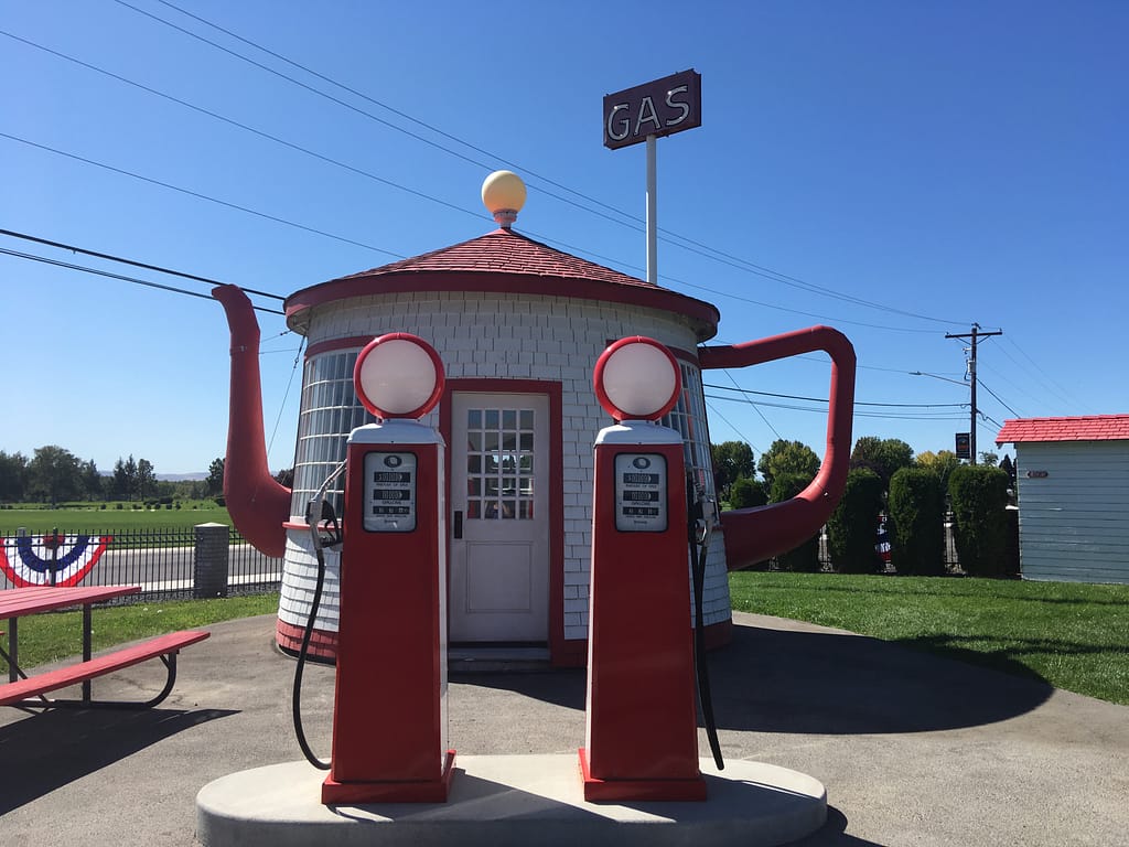 A very cute circular white building with a red roof, teapot handle and spout, with two vintage red gas pumps in the foreground.