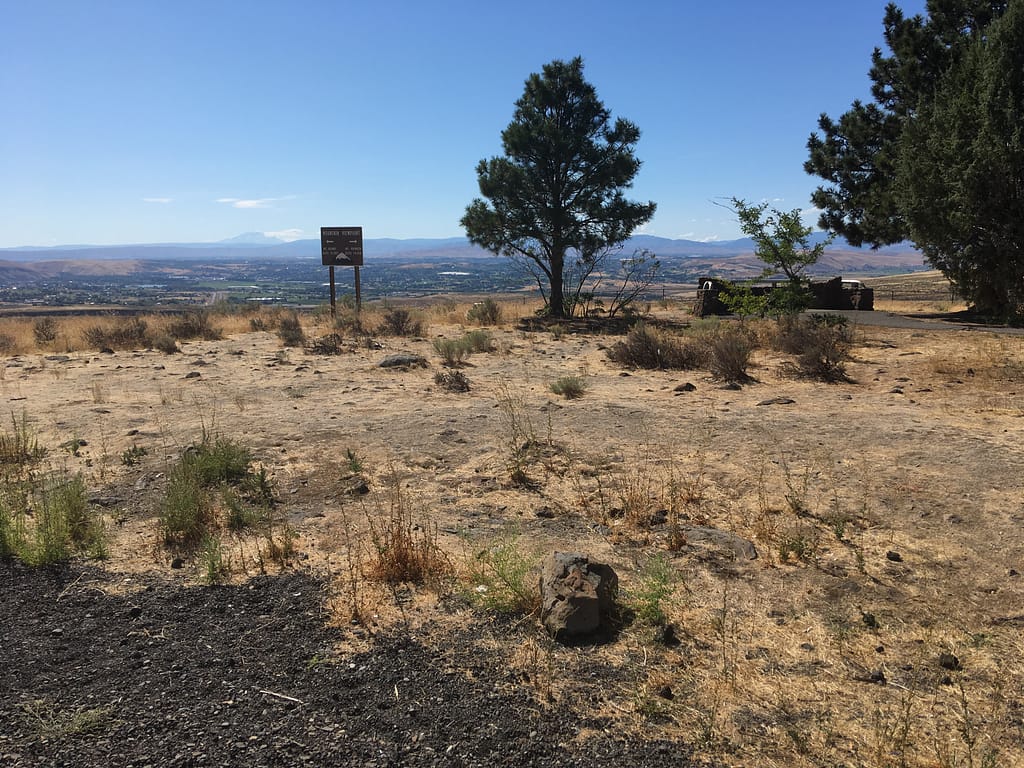 Scenic overlook in the Yakima Valley. A few pine trees, flat ground covered with golden-brown dry grass, and a sign in the distance pointing out the peaks of Mt Adams and Mt Rainier.