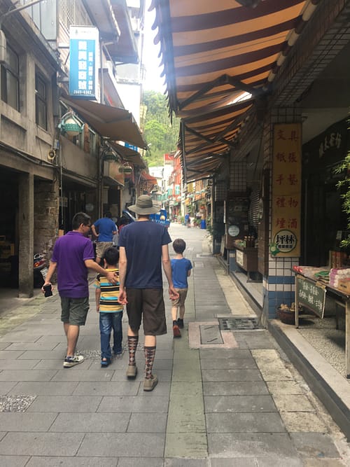 Four people walking through a narrow alleyway with small shops on either side.