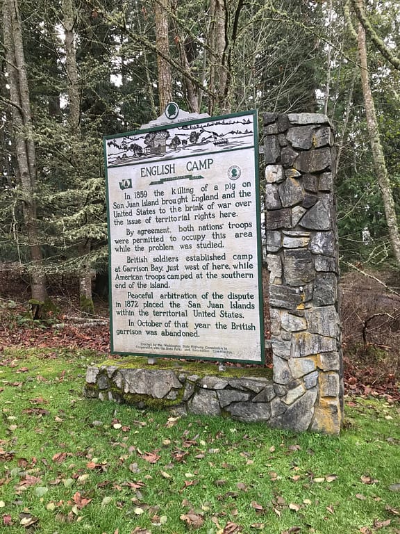 Sign reads, "English Camp. In 1859 the killing of a pig on San Juan Island brough England and the United States to the brink of war over the issue of territorial rights here. By agreement, both nations' troops were permitted to occupy this area while the problem was studied. British soldiers established camp at Garrison Bay, just west of here, while American troops camped at the southern end of the island. Peaceful arbitration of the dispute in 1872 placed the San Juan Islands within the territorial United States. In October of that year the British garrison was abandoned."