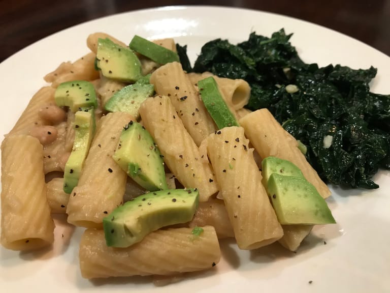 Rigatoni pasta mixed with white bean sauce and topped with sliced avocado and black pepper, with a side of braised kale