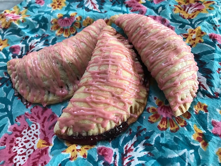 three cherry turnovers with pink icing arrayed on top of a blue floral cloth